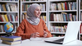 Muslim female teacher in hijab giving video call lesson using laptop in campus library space. The tutor or coach has remote e-learning for students, online training, conducts a course, talks remotely