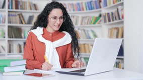 Female student on online e-learning taking notes or video calling remote teacher using laptop computer in campus library space. Brunette woman applicant is preparing for university exams in classroom