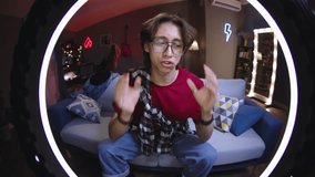 Asian guy with glasses shoots conversational emotional stream while sitting on sofa in front of ring led lamp. Recording video for likes and followers on social media platforms. Virtual life concept