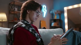 Asian guy in glasses with tablet sits on the couch, online communication. Virtual life in social media. Scrolling pages in social networks with friends, followers, likes, haters, reading book, news