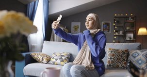 Happy modern Muslim woman in hijab taking selfie with front camera of phone. Middle plan of islamic girl taking selfie photo in modern interior. Attractive woman in hijab looking to phone camera 