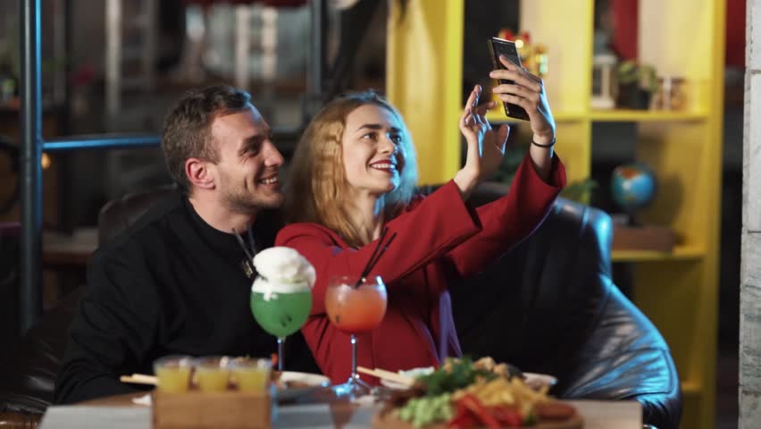 The boy and the girl are sitting in a restaurant, they are in a good mood. The girl holds a phone in her hands and tries to take a selfie, and they pose together.