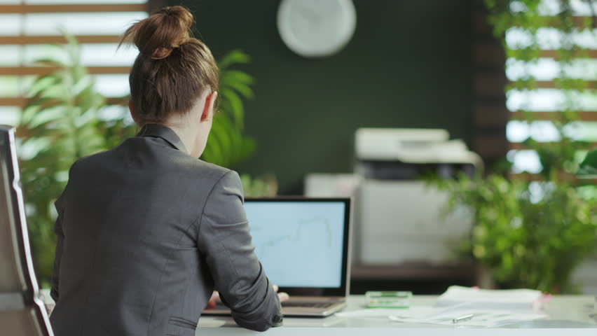 Sustainable workplace. Seen from behind modern small business owner woman in a grey business suit in modern green office stretching hand.
