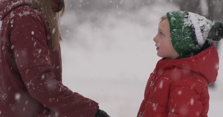 Mom straightens her sons hat and clothes and kisses him on nose outside when it snows. Mother or nanny takes care of child while walking in snow-covered park. Royalty-Free Stock Footage #1107671399
