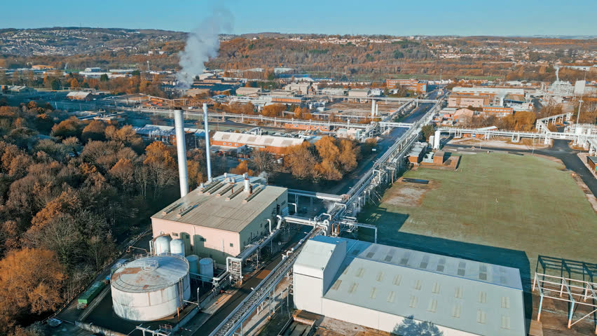 Aerial footage moving towards a large industrial chemical plant, in the uk. Showing pipelines, metal structures, cooling towers with steam and chemical storage tanks.