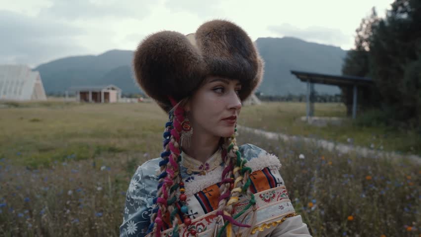 A Close-Up Shot of a Young European Female in Traditional Tibetan Clothing Standing in the Middle of a Green Meadow with Yellow Flowers and Beautiful Mountains in the Background 4K Footage. 