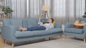 Full Body Of Asian Teen Girl Having Video Call On Smartphone While Lying On Sofa In The Living Room At Home. Waving Hands, Smiling, And Speaking
