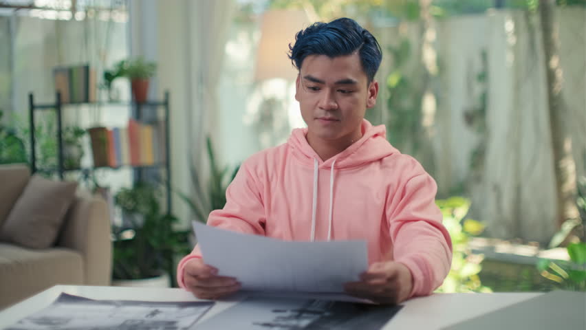 Portrait of young man with blue hair in pink hoodie showing black and white photograph he made to camera while sitting at table