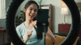 Beautiful young asian woman using mobile phone recording, live streaming a video vlog sharing on social media. Asian female blogger online live streaming, dancing sitting on sofa at home.