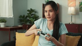 Excited beautiful Asian woman using joystick wireless controller enjoy playing fighting console video game to win highest victory. Girl Gamer sit on sofa in modern living room at home