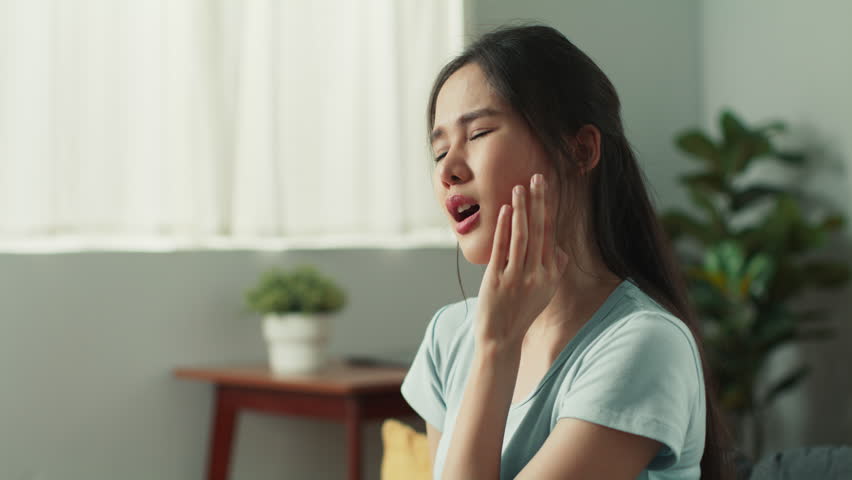 Attractive young asian woman touching cheek suffering from tooth pain sitting on couch at home. Distressed Asian woman having severe toothache feel unhealthy and unwell sit in living room at home. Royalty-Free Stock Footage #1107684121