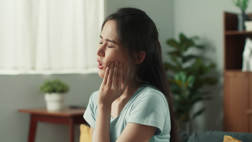 Attractive young asian woman touching cheek suffering from tooth pain sitting on couch at home. Distressed Asian woman having severe toothache feel unhealthy and unwell sit in living room at home. Royalty-Free Stock Footage #1107684133