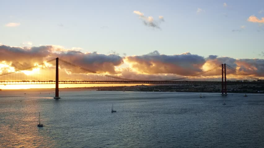 View of 25 de Abril Bridge famous tourist landmark of Lisbon connecting Lisboa and Almada on Setubal Peninsula over Tagus river with tourist yacht boats on sunset. Lisbon, Portugal