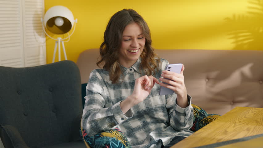 Positive girl laughing with smartphone in the hand. Joyful student resting at home. Smiling woman chatting online on mobile phone. Cheerful lady with happy face expression. Digital concept, 4k footage