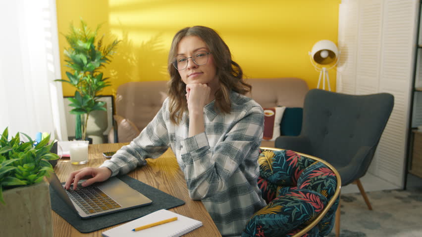 Confident woman working at remote workplace. Smart girl typing on laptop keyboard at home. Collage student learning online. Serious lady posing on camera. Female model holding hand on the chin