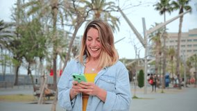 Slow motion fotage of one young adult woman walking and having fun using a cellphone social media application. Blonde feamale texting and sending funny messages on her smartphone. Lady holding phone