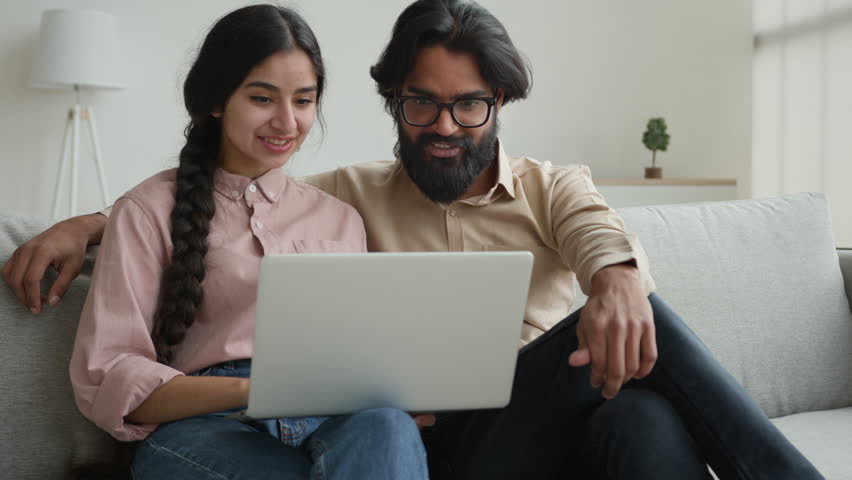 Arabian Indian couple on couch use laptop computer shopping online together pay bills order food at home choose buying internet goods give high five gesture happy man and woman approve teamwork unity Royalty-Free Stock Footage #1107695447