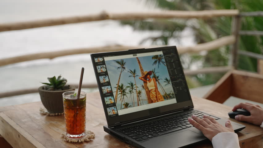 Female photo editor working with image editing software. Travel photographer edits photos. Retoucher freelancer woman doing color correction for pictures on laptop screen at tropical restaurant.