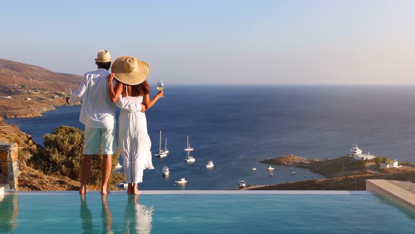 A hugging, loving couple on vacation time enjoys the summer sunset over the Aegean Sea by the swimming pool with a drink
