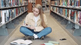 Smart girl student sitting on floor among bookshelves in university campus library learning, schoolgirl doing college course work study, doing research, writing notes reading book.
