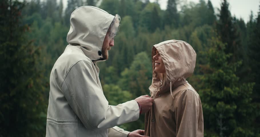 The guy fastens the girl's jacket and kisses her during the rain in a mountain coniferous forest. Happy couple against green forest background Royalty-Free Stock Footage #1107715473