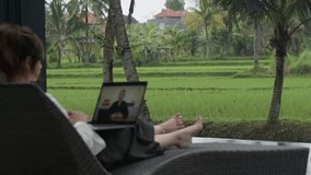 Rear view of young woman sitting on a lounge chair, watching a video on laptop. Panoramic view on tropics. Concept of online education, traveling and freelance