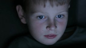 A boy watches a cartoon on a laptop or tablet in a dark room. The light from the screen plays on the face of a pretty boy about ten years old.