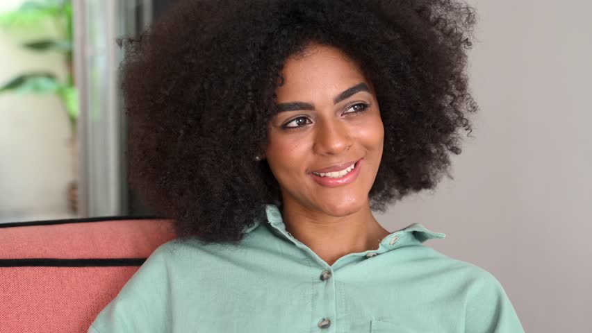Young African American female with curly hairstyle and beautiful smile looking aside sitting on the sofa with smartphone in hands, blurred home interior on the background, satisfied and pleased | Shutterstock HD Video #1107718985