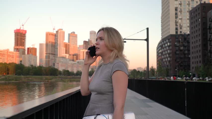 A young woman stands by the river and talks on the phone against the backdrop of skyscrapers