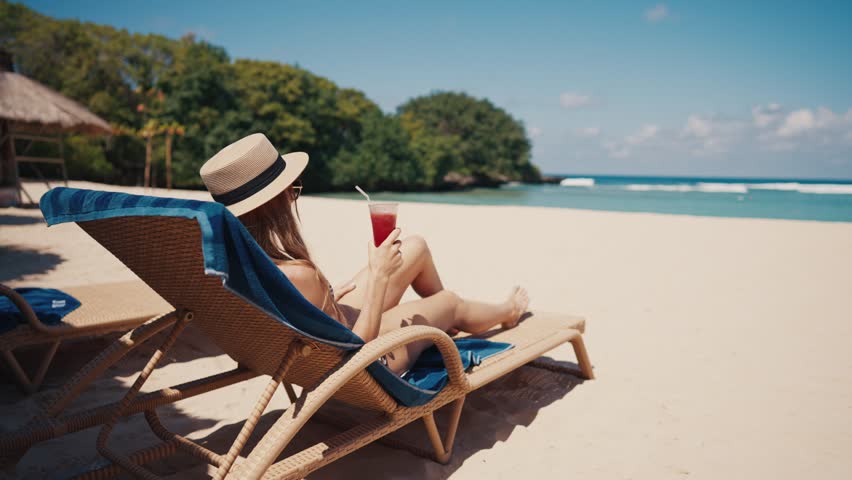 Relaxed woman in sun hat lying in sunbed on sandy ocean beach drinking fresh cocktail looking at turquoise ocean water. Summer vacation, beach resting, enjoying tanning. Travel, tourism, journey. Royalty-Free Stock Footage #1107722195