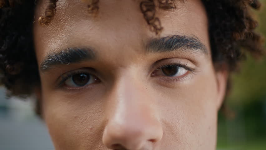 Brown eyes man looking camera outdoors extreme closeup. Curly hair teen staring lens attentively at street portrait. Handsome youngster clear sight detailed macro view. Focused human face expression