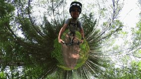 Goofy looking 360 type tiny-planet clip of a man riding a mountain bike along a muddy trail that is surrounded by trees.  The distorted view makes the man’s head very large.
