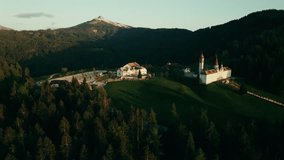 The drone flies around a monastery with a chapel standing among a dense spruce forest on top of a mountain. Drone video at sunset of a summer day