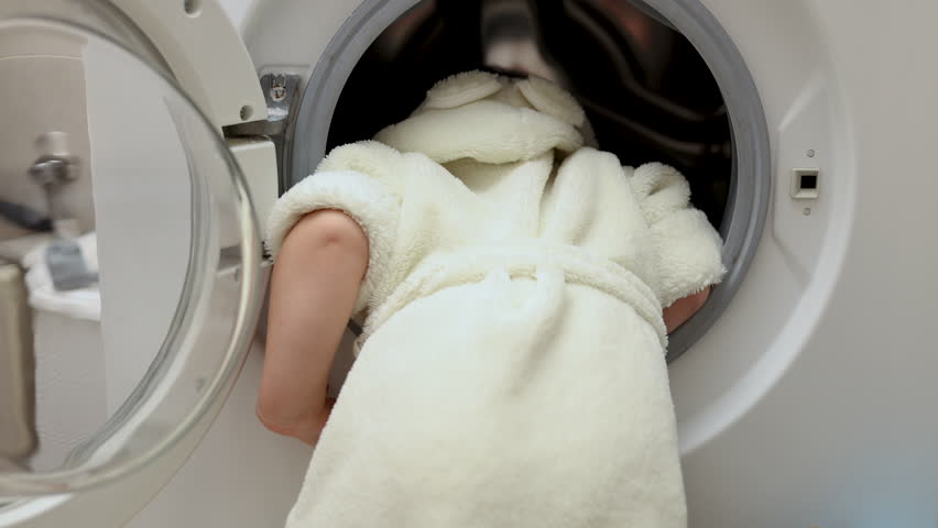 adorable cute baby boy in bathrobe putting or remove clothes from washing machine.kid playing with glass door.funny face emotions,top side view,preschooler child.4k bathroom interior.