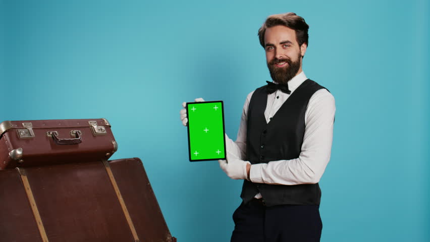 Elegant bellboy holding tablet on camera and presenting blank greenscreen display, symbolizing hotel concierge services with professionalism. Porter in suit and tie pointing at isolated mockup. Royalty-Free Stock Footage #1107753427
