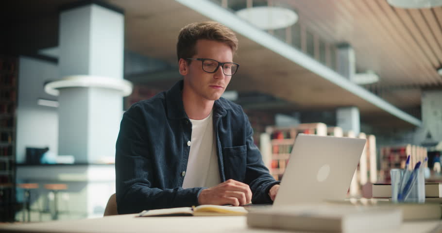 Young Specialist Working in a Technology Startup Company. Handsome Man Taking University Lessons After Work, Studying in Campus Library. He is Using Laptop Computer, Writing a Homework Project Online