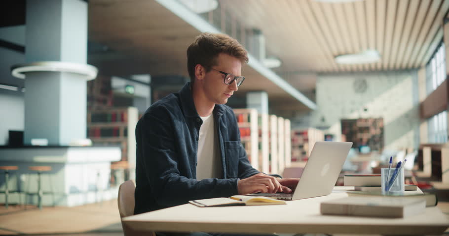 Handsome Male Student Working on His University Homework on a Laptop Computer in a Modern Library. Young Caucasian Man Looking at Camera, Smiling. He's Wearing Glasses and a Dark Shirt Royalty-Free Stock Footage #1107760491