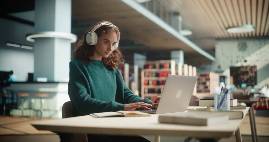 Happy and Focused Student Studying in a Modern Library. Young Female Wearing Headphones for Comfort and Silence, Using Laptop Computer to Work on a University Research Project Online Royalty-Free Stock Footage #1107760503