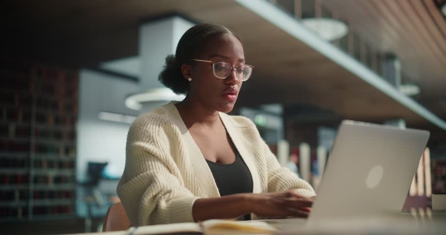 Portrait of a Stylish African Female Student in Glasses Working on Her University Exercise on a Laptop Computer. Young Black Woman Studying an Online Course in an Empty Quiet Public Library