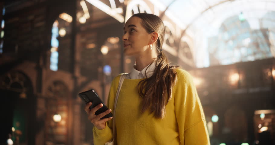 Casual Female Student Walking in an Public Library with Historical Wooden Interior. Young Smart Woman Looking Around, Using a Smartphone, Chatting with Friends Online, Browsing Social Media Royalty-Free Stock Footage #1107760531