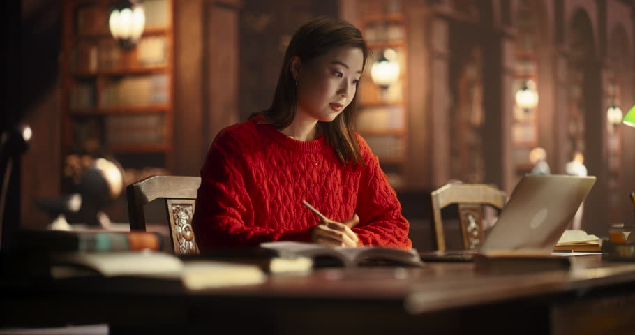 Attractive Multiethnic Student Making a STEM Class Homework in a Textbook, While Researching Quiz Answers on a Laptop Computer. Young Asian Female Studying in a Public Library with Classical Interior