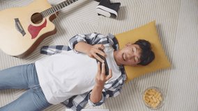 Top View Of Asian Teen Boy Enjoys Playing Game On Smartphone While Lying On Carpet On The Floor At Home

