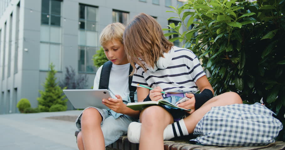 School students friends outdoors using digital tablet computer . Kids sitting on bench and play on laptop near school campus outdoors . Diverse kids doing a project together with tablet and notebook. | Shutterstock HD Video #1107763433