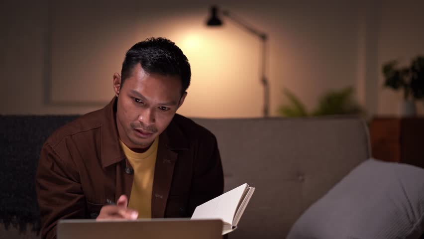Asian man freelancer study or learn in his laptop computer at  with notebook sitting on sofa at home Online e-learning at night room Royalty-Free Stock Footage #1107766457