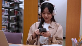 Young beautiful Asian woman student using smartphone to listening or searching lesson to studying while reading book at desk at university in college