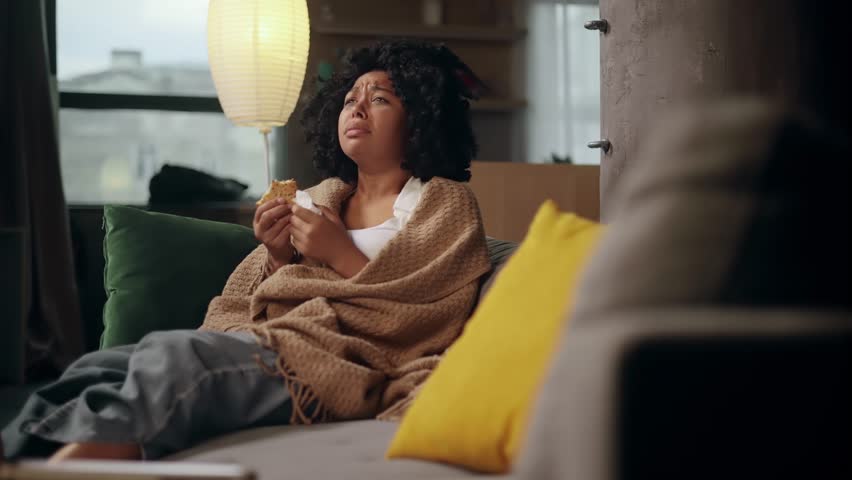 Portrait of sad young woman dealing with stress by eating food sitting on sofa at home Upset female wrapped in blanket crying and eating cookies indoors alone Mental heath problem Royalty-Free Stock Footage #1107767667