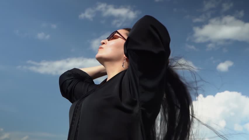 a girl in a black long dress with long hair fluttering in the wind stands against a blue sky with clouds