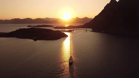 Stunning view from above of a sailboat at sunset, midnight sun, in Lofoten, Norway, with mountains in the distance, yellow and orange sky. 4K resolution, 30fps.