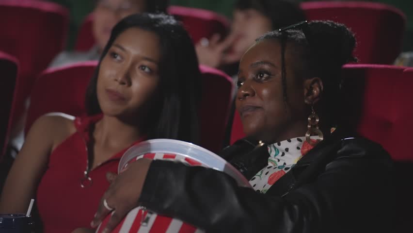 Two young women enjoy watching a movie in theater. Female friends sharing popcorn while watching a movie in the cinema. Two young woman friends laughing happily while watching movie in cinema hall Royalty-Free Stock Footage #1107791135