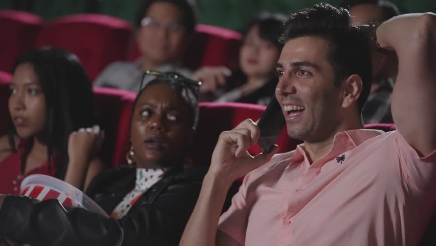 Young hispanic latin man talking on smartphone while watching movie at the cinema and disturbing audience. Phone talks prohibited during movie show. A man disrespectful talking on phone at theater